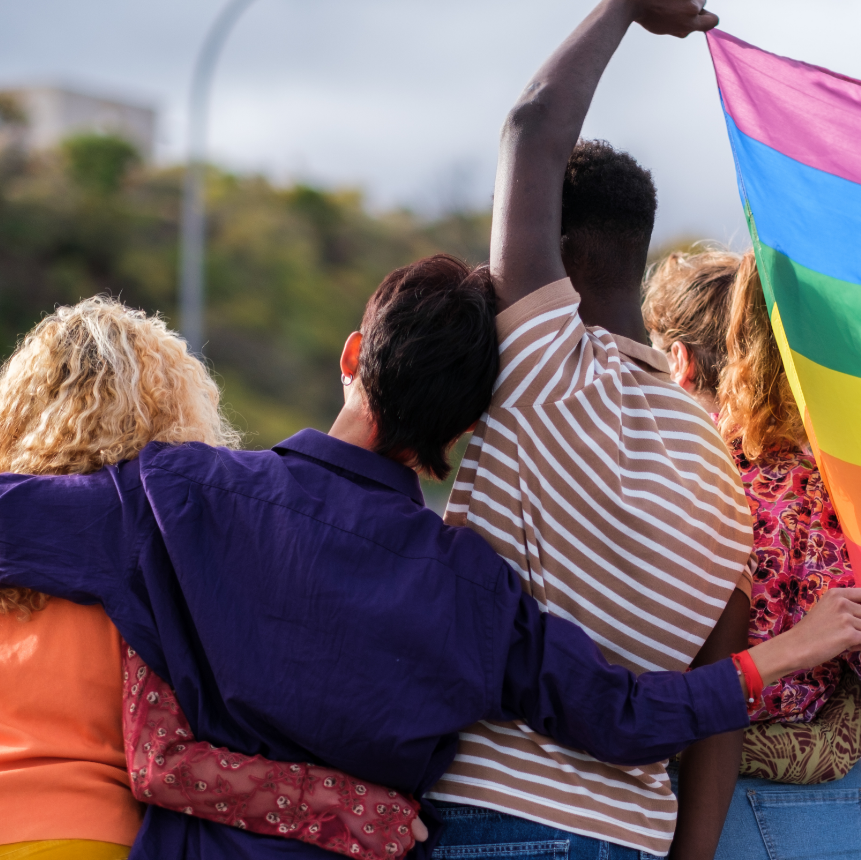 group of diverse youth, arm in arm and holding a pride flag with their backs to the camera.